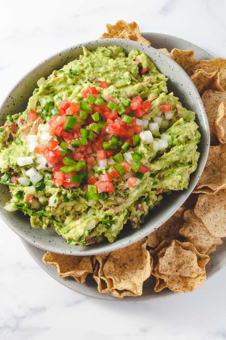 fresh guacamole in a gray bowl, on a gray plate with chips on the plate. onion tomatoes and jalapeños on top as garnish