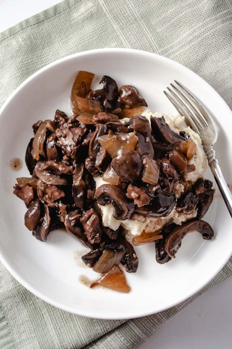 crockpot sirloin beef tips on a white plate with fork over mashed potatoes on a green towel.