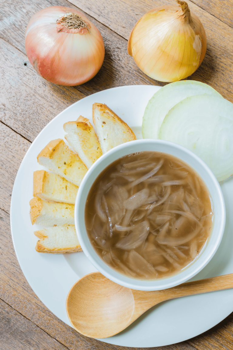 Easy Instant Pot French Onion Soup with Red Wine top down view of Easy Instant Pot French Onion Soup in a white bowl on a white plate with onions and bread on the plate on a wood table. with a wooden spoon on plate