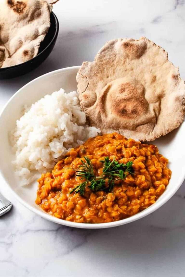 vegan red lentil curry in a white bowl with white rice and a naan bread on white counter with naan bread in a black bowl at the top left