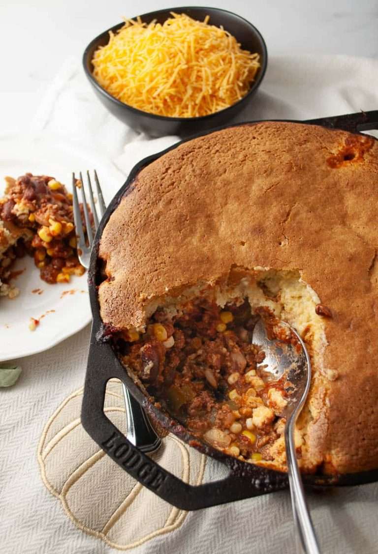 Easy Chili Cornbread Skillet Casserole Bake Recipe chili cornbread skillet bake on a lodge cast iron skillet on a cream and white cloth with a white plate with a serving with a fork and a black bowl with cheddar cheese in it to serve on top top view with a sering cut out of the skillet