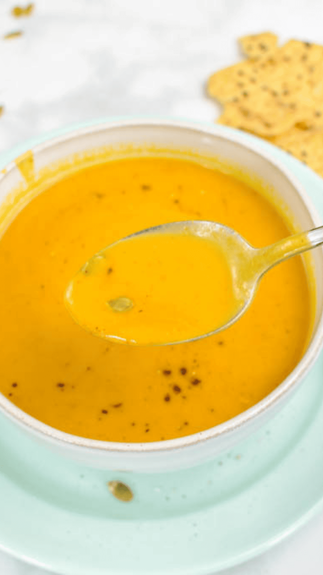 Pumpkin in a cream bowl on a white counter with pumpkin seeds on top and a spoon on the side on a green plate close up of the spoon with soup and crackers in the background