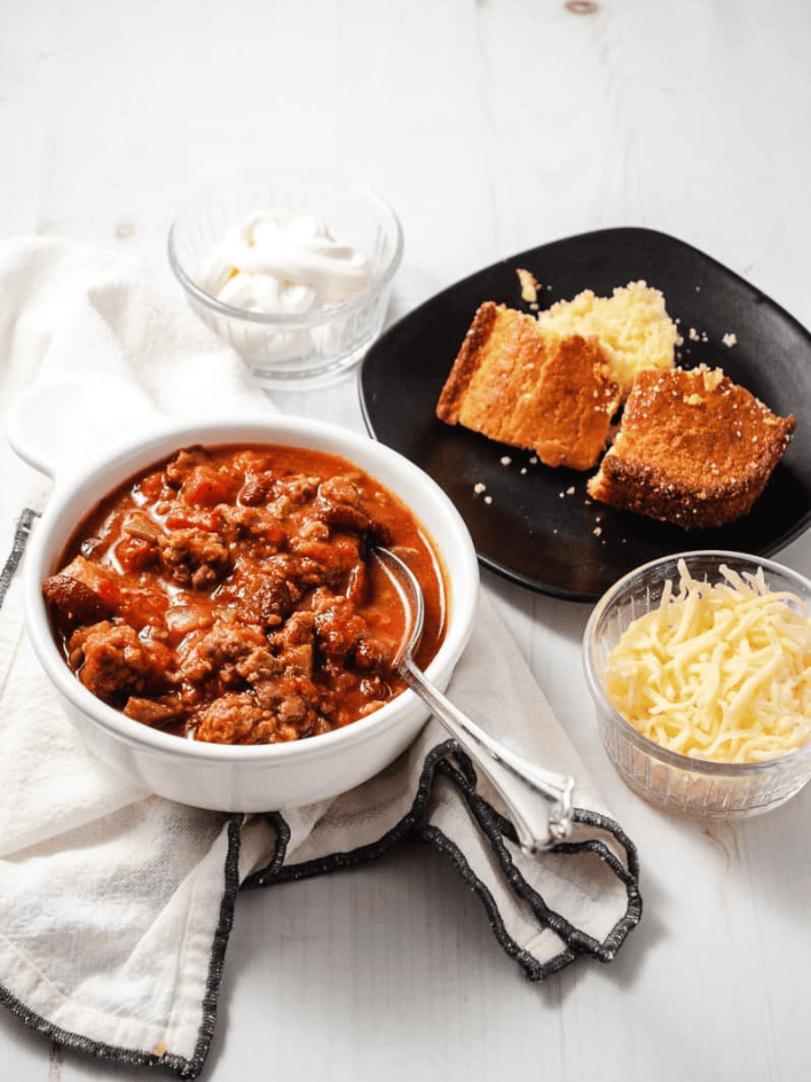 white bowl of chili with spoon on white napkin with black trim. on a white wood table. 2 clear bowls one sour cream one shredded cheese a black plate with 2 squares of corn bread.