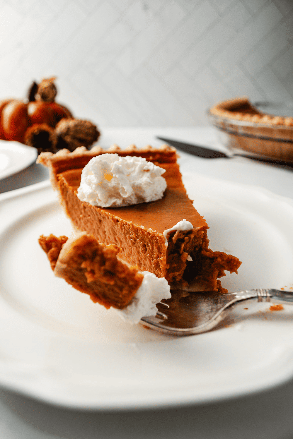A slice of Classic pumpkin pie on a white plate looking straight onto a fork with a bite and a fork whip cream on top of the pie the pie and serving knife pinecones and a glass pumpkin in the background