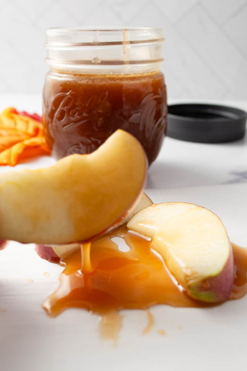 easy-maple-syrup-caramel-sauce in a small mason jar and an apple slice dripping wih sauce. 