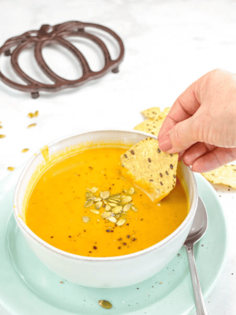 a white bowl of instant pot pumpkin soup on a green plate with a spoon a woman's hand Divina cracker into the soup