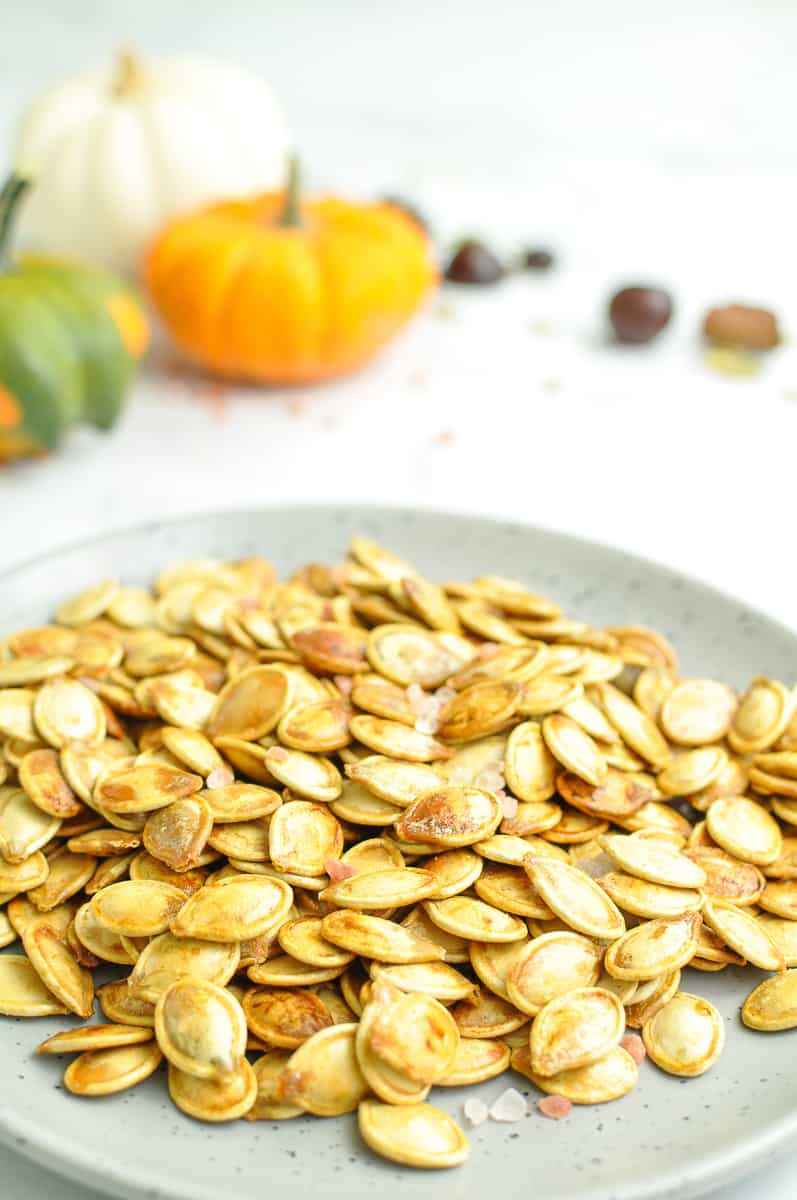 A plate filled with golden roasted pumpkin seeds sprinkled with coarse salt sits in the foreground, capturing the cozy essence of fall baking. In the softly blurred background, small decorative pumpkins in orange, white, and green add a warm autumn touch, making this an inviting scene for anyone looking for easy fall recipes or simple pumpkin snacks.