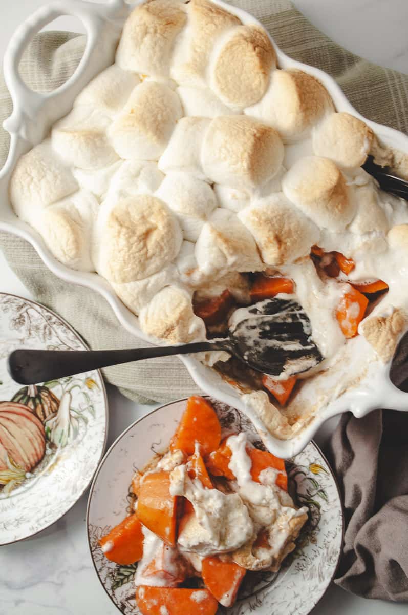 Candied sweet potato casserole in a casserole dish on a green towel with two Thanksgiving plates and a scoop of casserole on one plate.