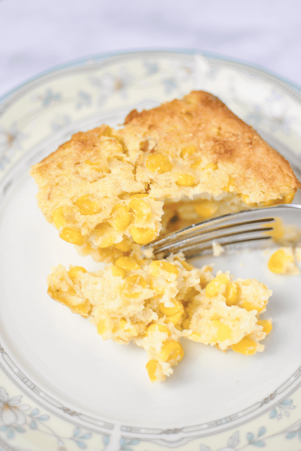 a slice of Easy Corn Bake  Casserole Recipe with Jiffy Mix on a white plant with a yellow and light blue flower border for taking a bite out of the casserole.