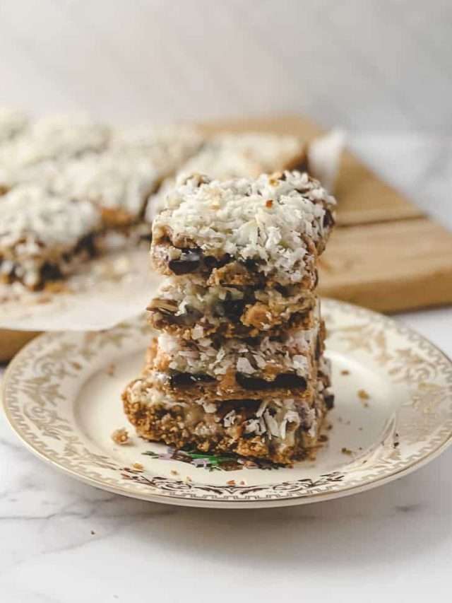 Easy 7 Layer Magic Cookie Bars stack of 5 squares on a gold flowered plate the 10 other cookie bars on parchment on a wood cutting board