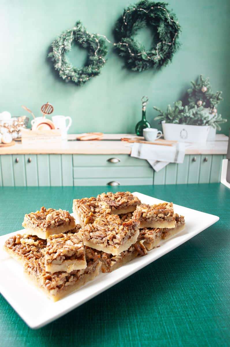 Easy Pecan Pie Shortbread Bar Cookie with Maple Syrup 9 cookie bars on a white rectangle tray on a green table cloth in a light green kitchen cabinets and butcher block counter in the background kitchen accessories on the counter showing more of the kitchen