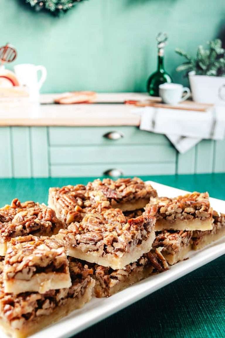 Easy Pecan Pie Shortbread Bar Cookie with Maple Syrup 9 cookie bars on a white rectangle tray on a green table cloth in a light green kitchen cabinets and butcher block counter in the background kitchen accessories on the counter.