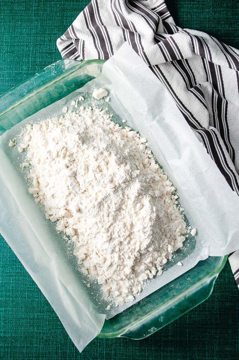 Rectangular baking pan lined with parchment paper with the butter flour and sugar mixture loose in the pan on the green counter with a navy and white striped towel.