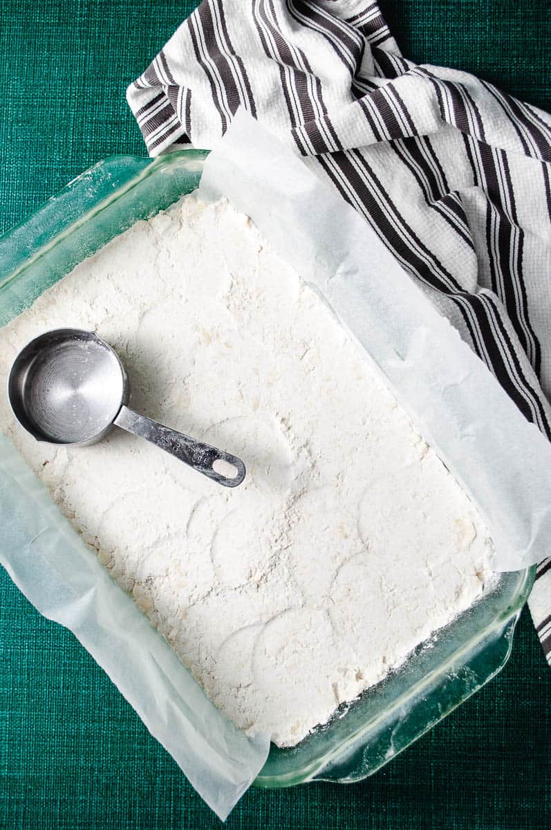 Particular baking pan lined with parchment paper with the flour mixture flour butter and sugar and a measuring spoon and a green counter with navy and white striped towel.