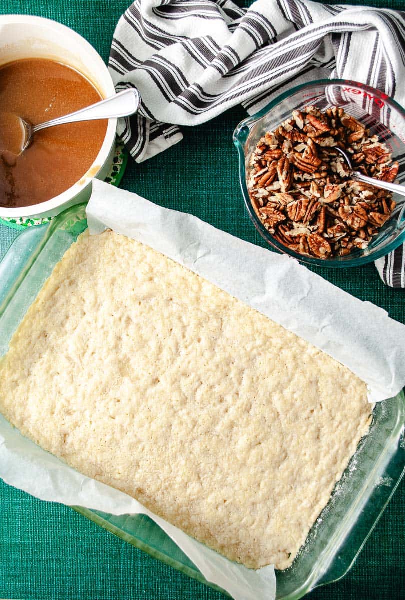 Baked shortbread crust in a rectangular baking pan with parchment paper line, with the chopped pecans in a measuring cup and the Maple syrup mixture in a pot with a spoon on the green counter waiting to be added to the crust.