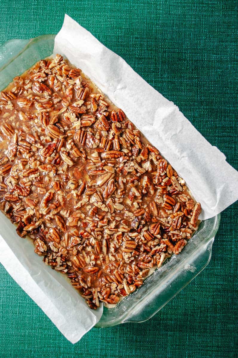 Rectangular baking pan lined with parchment paper with the pecan topping on top of the shortbread mixture on a green counter.