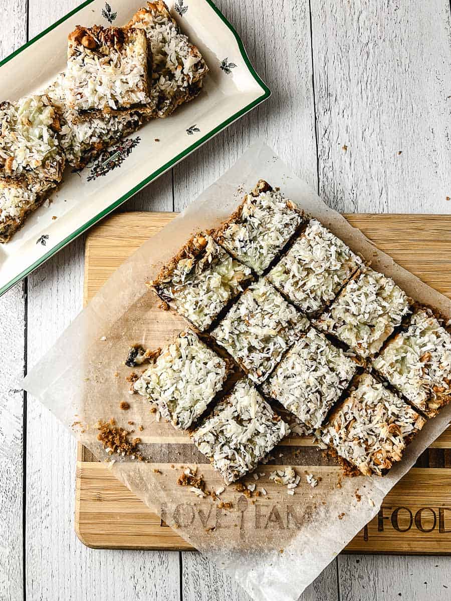 7 Easy 7 Layer Magic Cookie Bars on parchment on a wooden cutting board on a white washed wooden table with a platter of the cookies in the background,