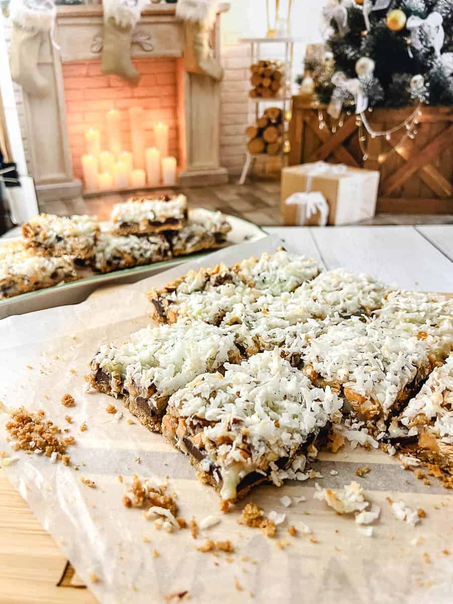 7 Easy 7 Layer Magic Cookie Bars on parchment on a wooden cutting board on a white washed wooden table with a platter of the cookies in the background, and a fireplace and Christmas tree in the background.