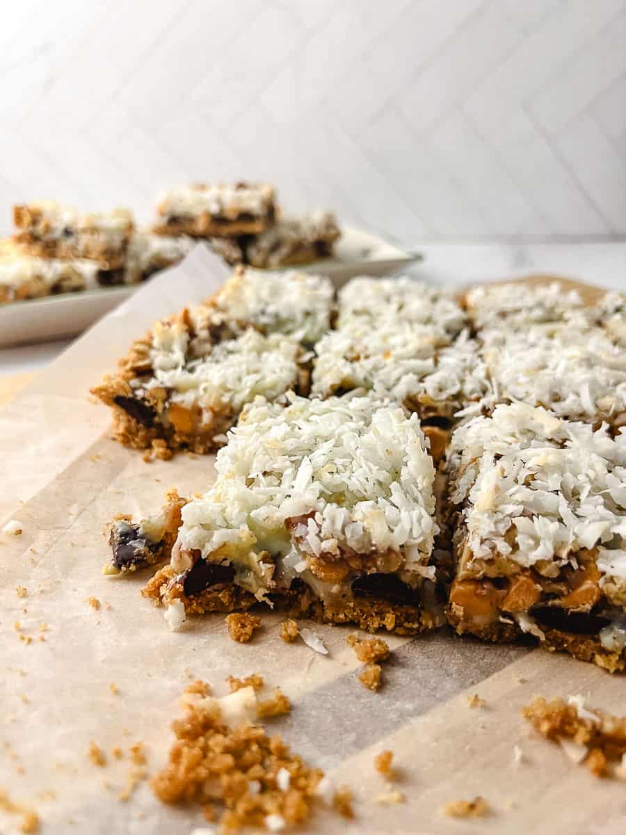 close up of the cut cookie bars 7 Easy 7 Layer Magic Cookie Bars on parchment on a wooden cutting board on a white washed wooden table with a platter of the cookies in the background, 