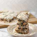 Easy 7 Layer Magic Cookie Bars stack of 5 squares on a gold flowered plate the 10 other cookie bars on parchment on a wood cutting board