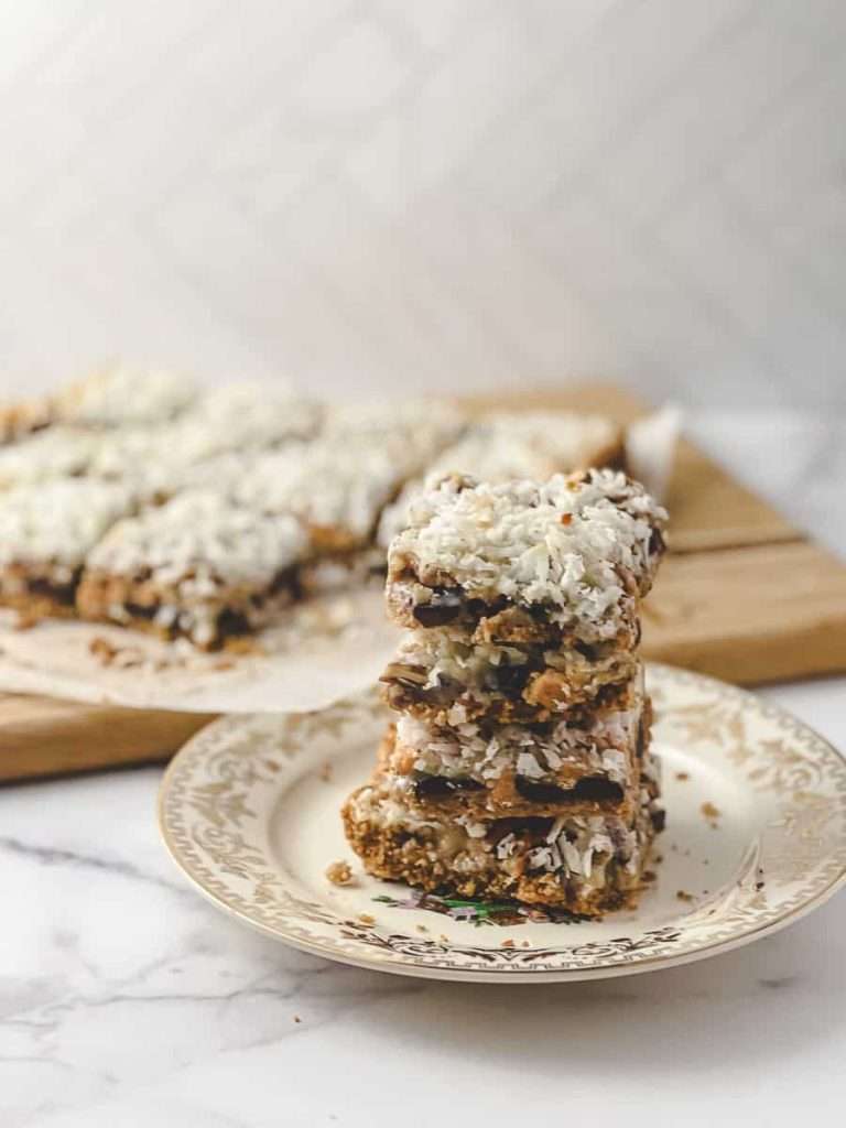 Easy 7 Layer Magic Cookie Bars stack of 5 squares on a gold flowered plate the 10 other cookie bars on parchment on a wood cutting board