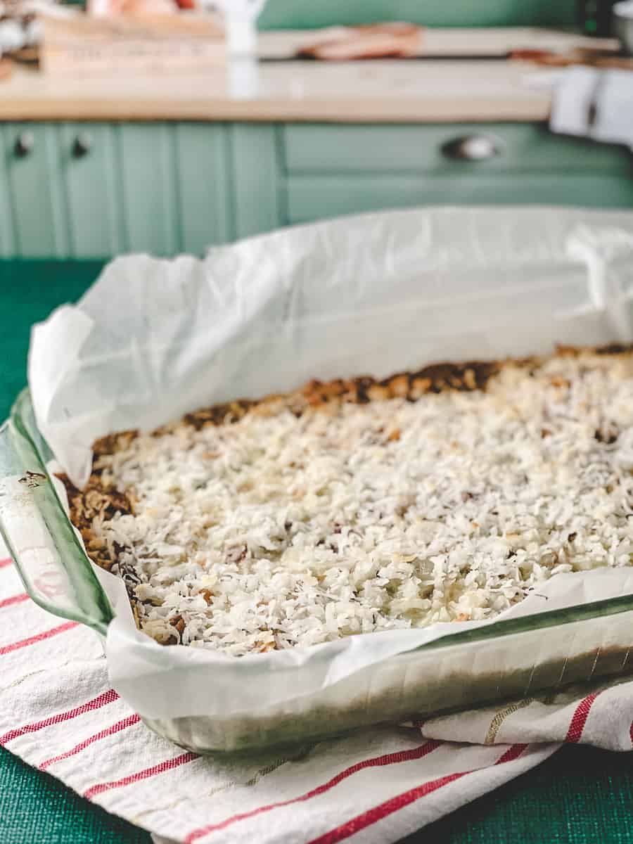 Easy 7 Layer Magic Cookie Bars Old Fashioned Favorite in a 13x9 baking pan lined with parchment on a red striped white kitchen towle.
