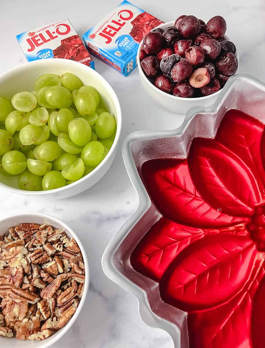 Best Christmas Jello Salad Mold Holiday Vintage Recipe red jello in pintsetta mold with a white bowl with grapes a white bowl with frozen red cherries and a white bowl with chopped pecans two red jello package on white counter top down view.