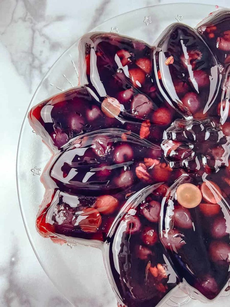 Classic jello mold recipe for Christmas, a close up of a deep red poinsettia shaped vintage jello mold showing grapes and cherries set inside the glossy jello on a clear glass plate.