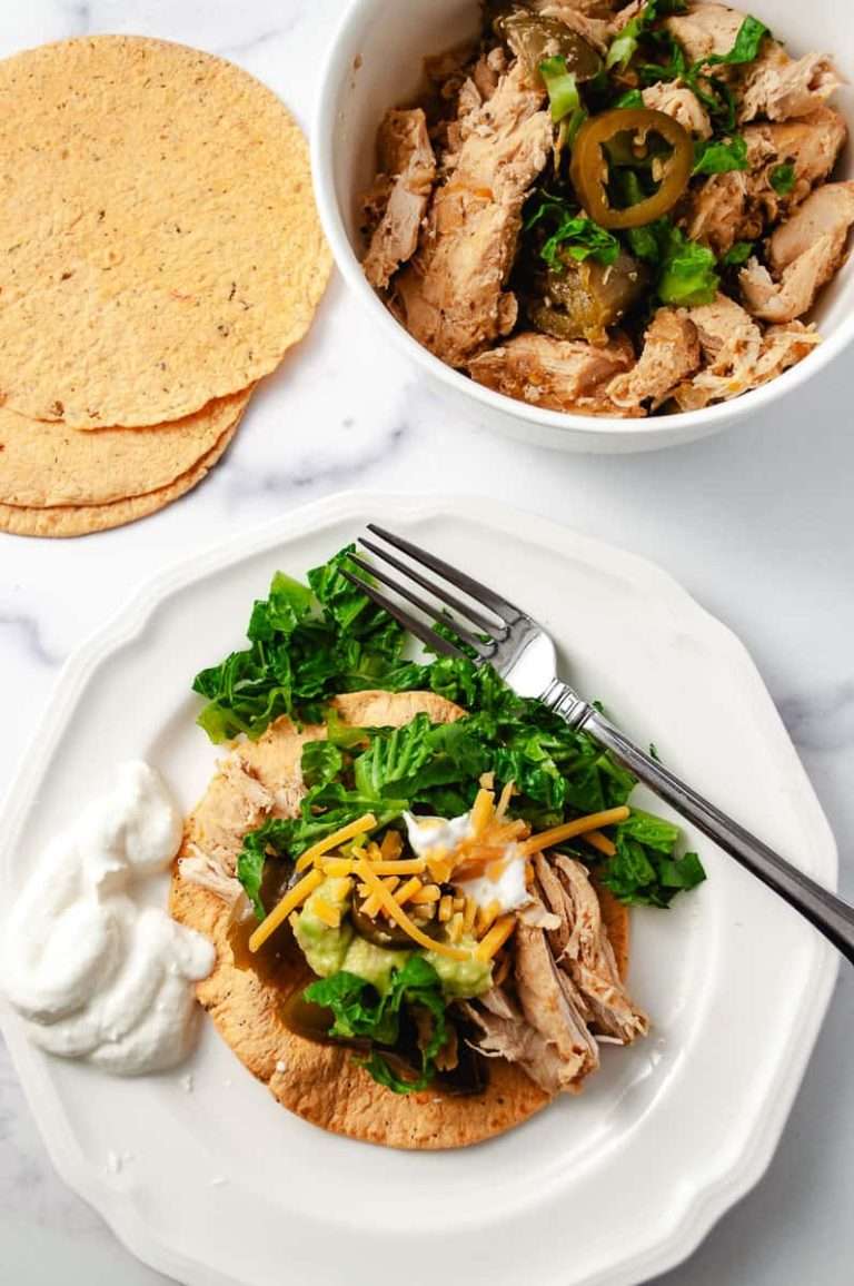 Instant pot juicy chicken fajitas easy recipe a serving on a corn tortilla with sour cream lettuce, shredded cheese and a fork on a white plate with corn tortillas on a white counter a a white serving bowl with the chicken fajitas