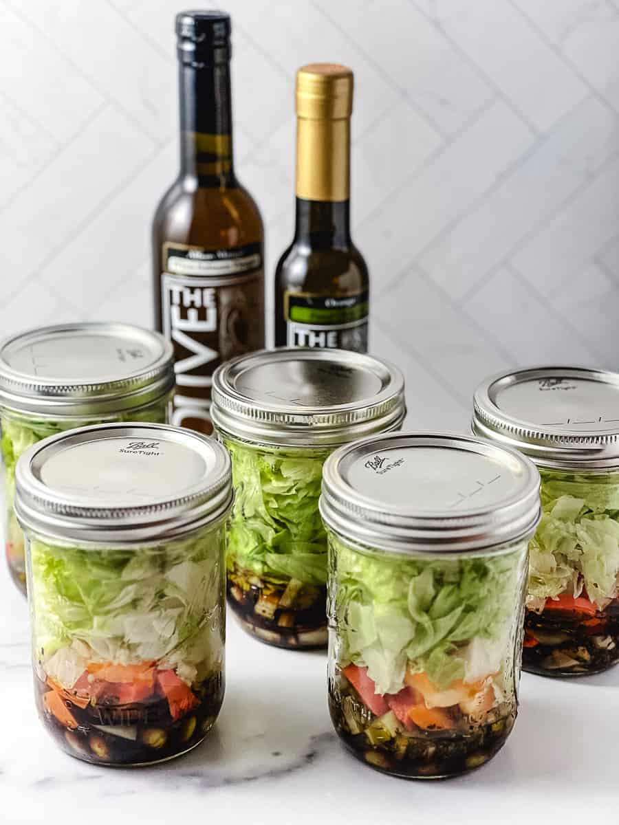 5 pint mason jars with salad layed in them with 2 jars one of vinegar and one of olive oil on a white counter