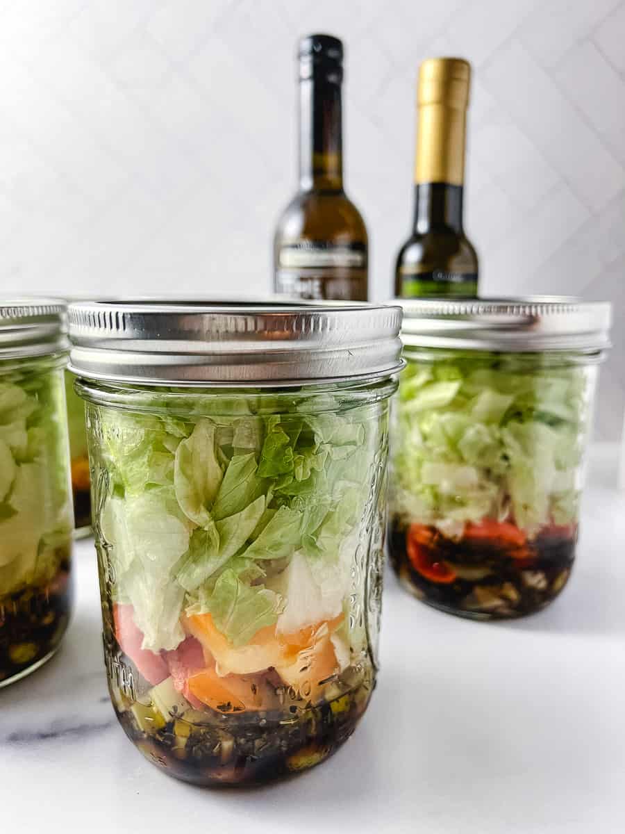 3 pint mason jars with salad layed in them with 2 jars one of vinegar and one of olive oil on a white counter