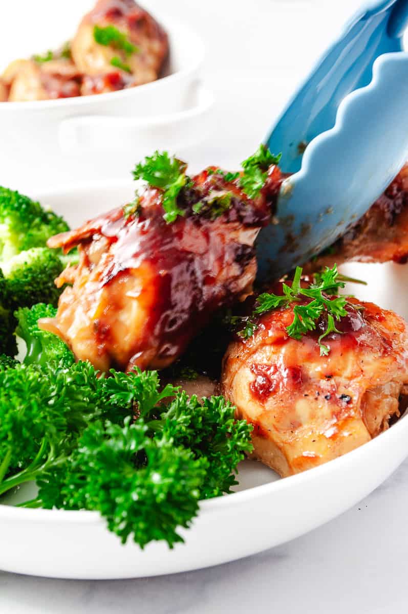 barbecue-chicken-drumsticks-crockpot-on a white plate with green parsley and tongs lifting up a drumstick. 