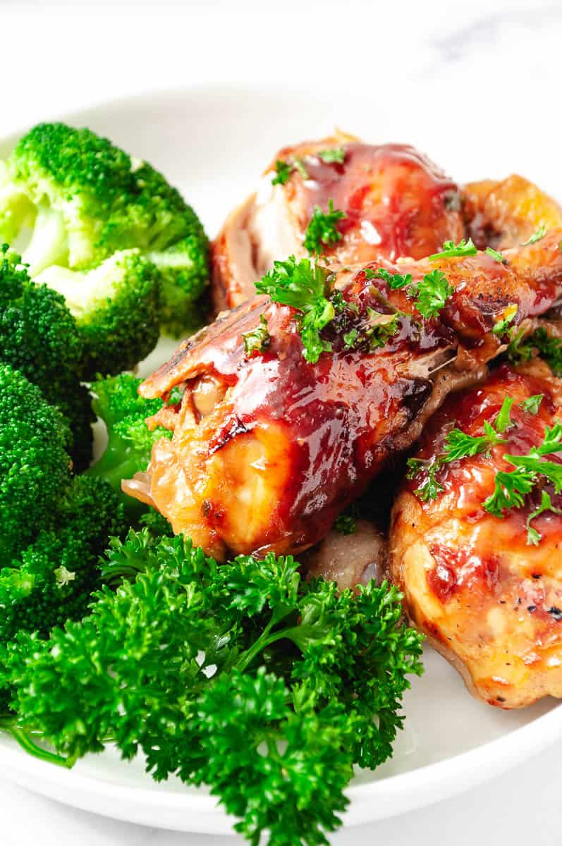 3 Ingredients BBQ Chicken Drumsticks CrockPot Recipe 3 drumsticks on a white plate with broccoli and parsley closeup.