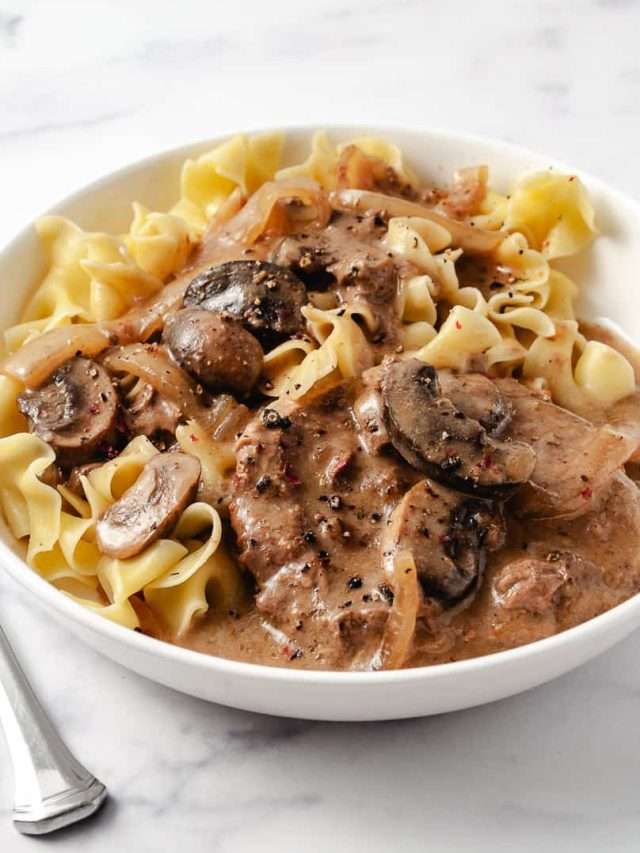 close up of Irresistibly Tender CrockPot Cube Steak with Gravy Recipe 2 meat patties with mushrooms and onions, over wide egg noodles in a white plate with a fork.