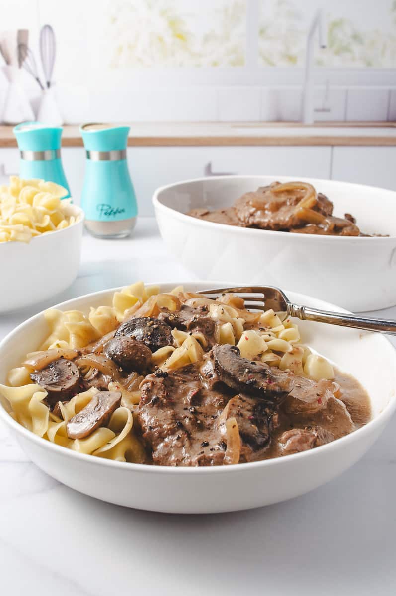 close up of the plate Irresistibly Tender CrockPot Cube Steak with Gravy Recipe 2 meat patties with mushrooms and onions, over wide egg noodles in a white plate with a fork on a white kitchen counter with a white bowl of egg noodles to the left, the serving bowl of cube steak to the right and aqua salt and peppers shakers at the top.