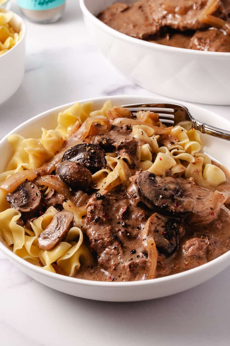 Irresistibly Tender CrockPot Cube Steak with Gravy Recipe 2 meat patties with mushrooms and onions, over wide egg noodles in a white plate with a fork