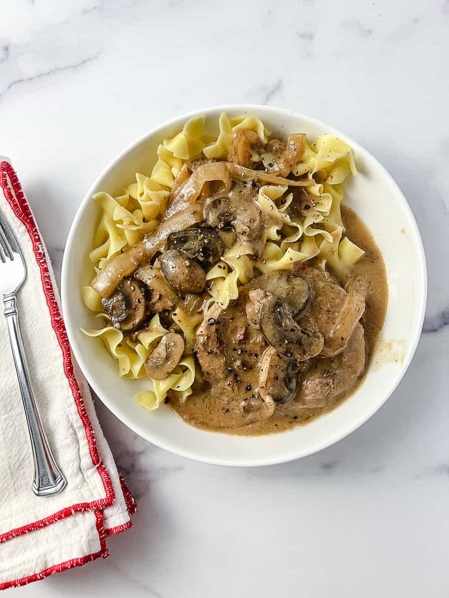 top down view Irresistibly Tender CrockPot Cube Steak with Gravy Recipe 2 meat patties with mushrooms and onions, over wide egg noodles in a white plate with a fork on a white napkin with red trim.