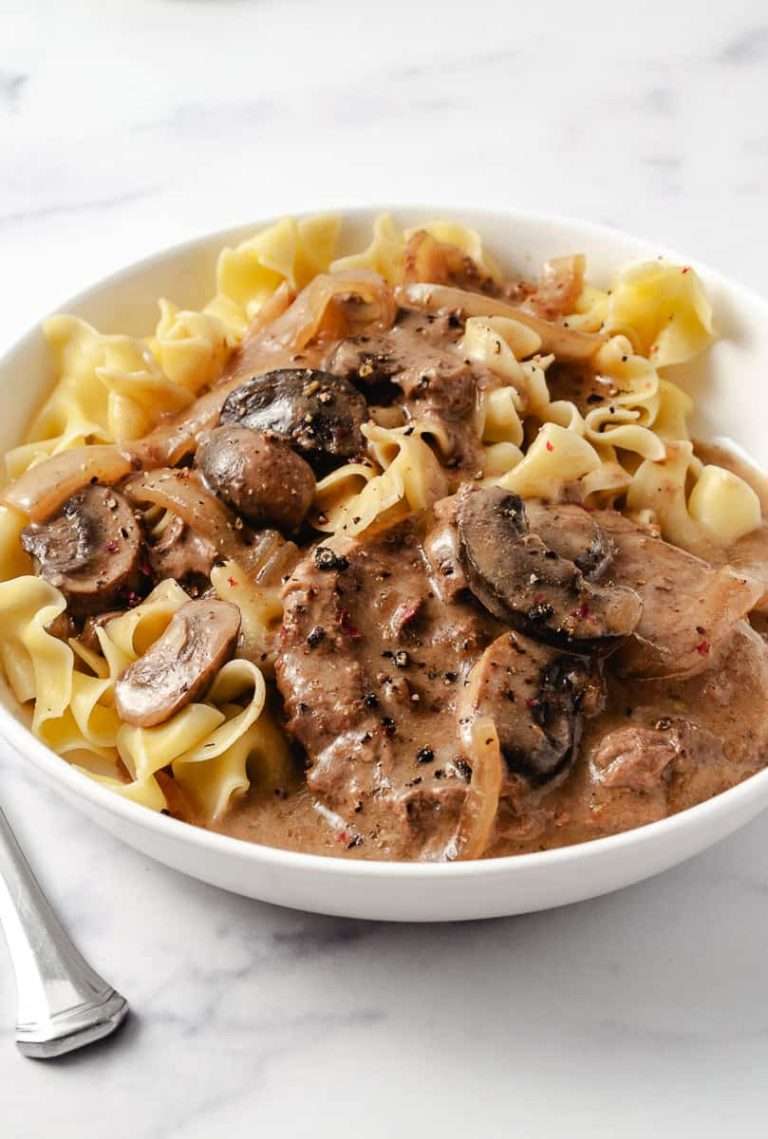 close up of Irresistibly Tender CrockPot Cube Steak with Gravy Recipe 2 meat patties with mushrooms and onions, over wide egg noodles in a white plate with a fork.