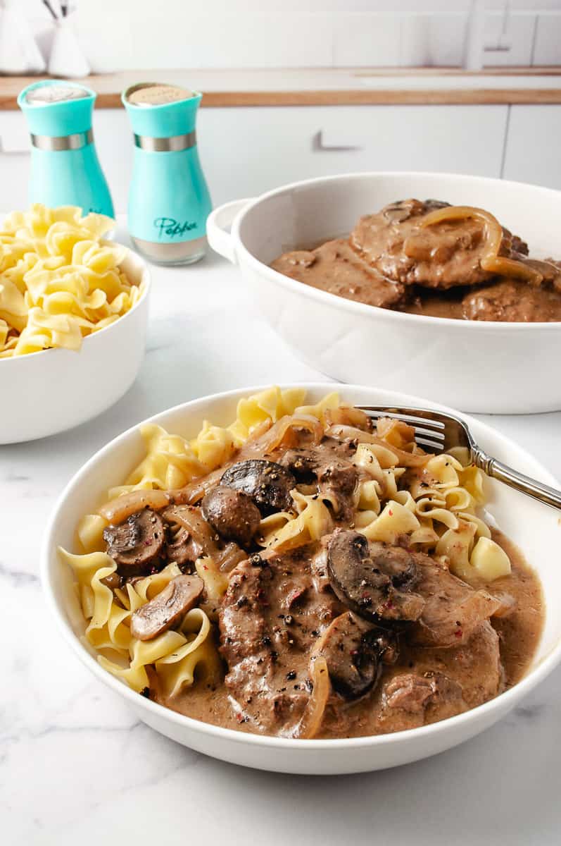 Irresistibly Tender CrockPot Cube Steak with Gravy Recipe 2 meat patties with mushrooms and onions, over wide egg noodles in a white plate with a fork on a white kitchen counter with a white bowl of egg noodles to the left, the serving bowl of cube steak to the right and aqua salt and peppers shakers at the top.