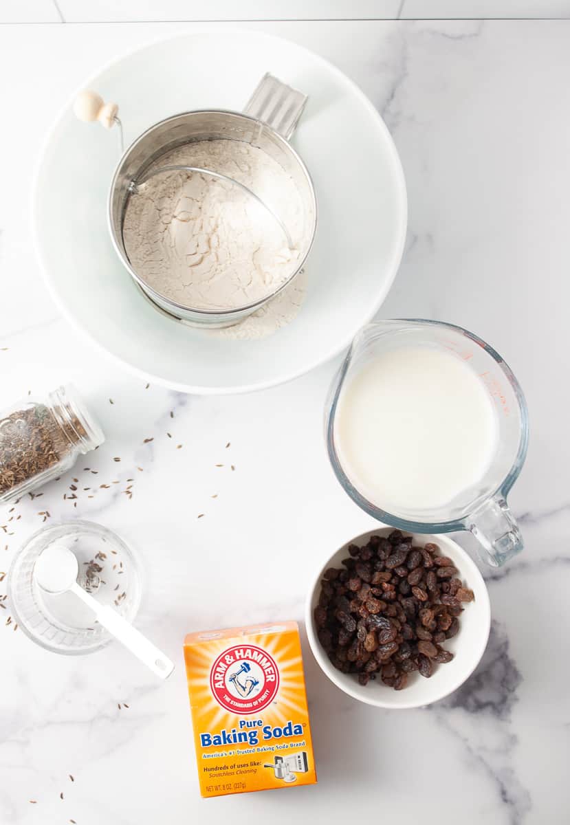 Irish Soda Bread Recipe Easy Knead with Bread Machine, top down view of ingredients on white counter flour in a sifter buttermilk in a glass pitcher raisins and a bowl baking soda box salt in a spoon and a clear bowl and caraway seeds in a spice jar and on the counter.