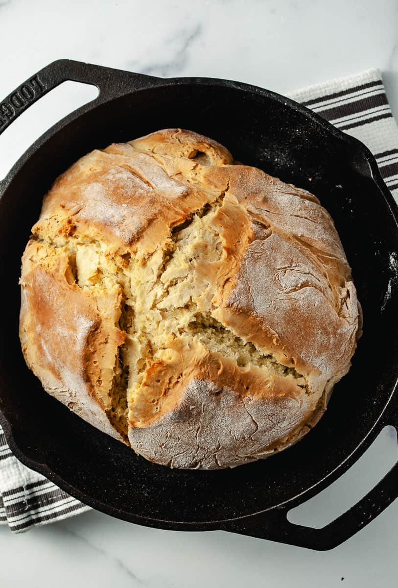 Irish Soda Bread Recipe Easy Knead with Bread Machine, golden brown load in cast iron pan on a gray and white striped towel. top down view