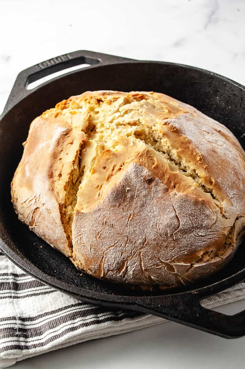 Irish Soda Bread Recipe Easy Knead with Bread Machine, golden brown load in cast iron pan on a gray and white striped towel.