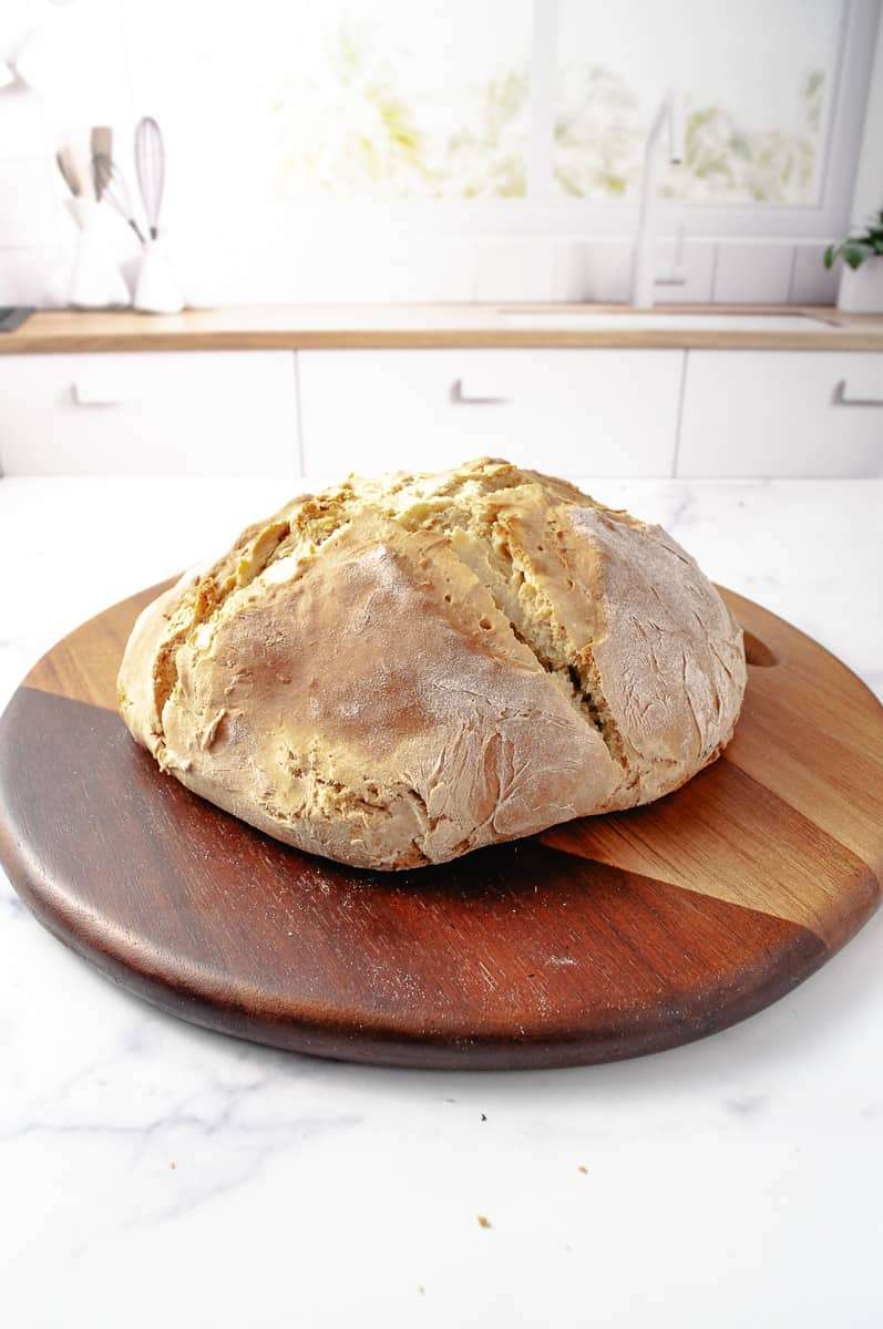 Irish Soda Bread Recipe Easy Knead with Bread Machine baked loaf on a wooden cutting board and a white counter in a white kitchen.