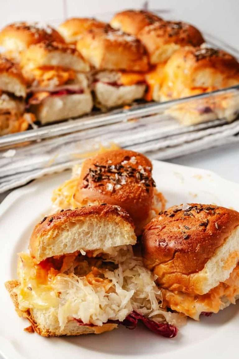 Reuben Sliders Sandwiches: Perfect Party Appetizer 3 sliders on a white plate the 13 by 9 pan in the background with the remaining sliders in a white kitchen.