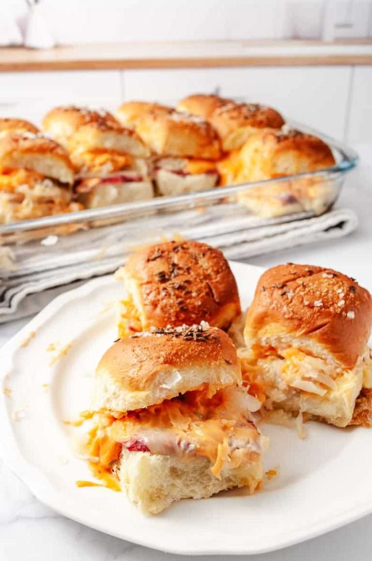 Reuben Sliders Sandwiches: Perfect Party Appetizer 3 sliders on a white plate the 13 by 9 pan in the background with the remaining sliders in a white kitchen.