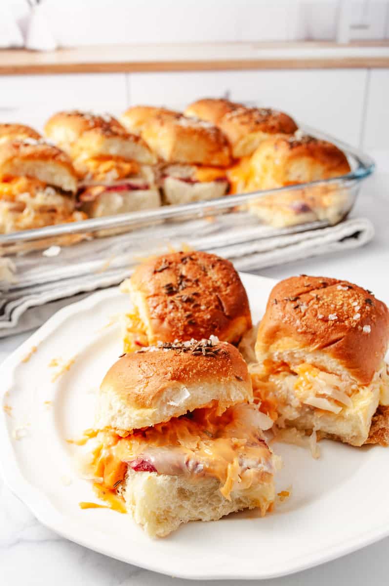 Reuben Sliders Sandwiches: Perfect Party Appetizer 3 sliders on a white plate the 13 by 9 pan in the background with the remaining sliders in a white kitchen.