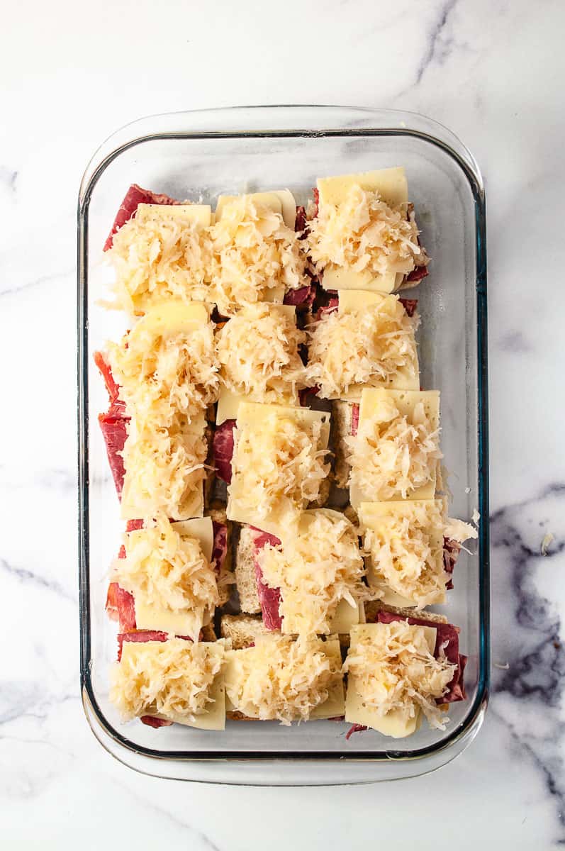 Reuben Sliders Sandwiches: Perfect Party Appetizer 15 sliders in a 13 by 9 baking pan with the corned beef, swiss cheese, sauerkraut on a white counter.