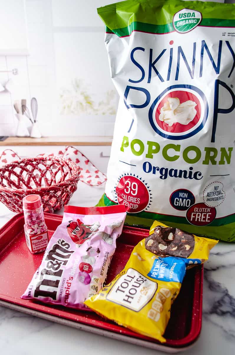 Easy Valentine's White Chocolate M&M Popcorn Treat ingredient on the counter with 2 trays one red and one pink on top of each other, m&ms, white chocolate chips, heart sprinkles skinny pop and a red ceramic basket bowl.