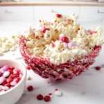 Easy Valentine's White Chocolate M&M Popcorn Treat in a red basket bowl on a white counter in a white kitchen with popcorn on the counter and a small bowl with M and M in it.