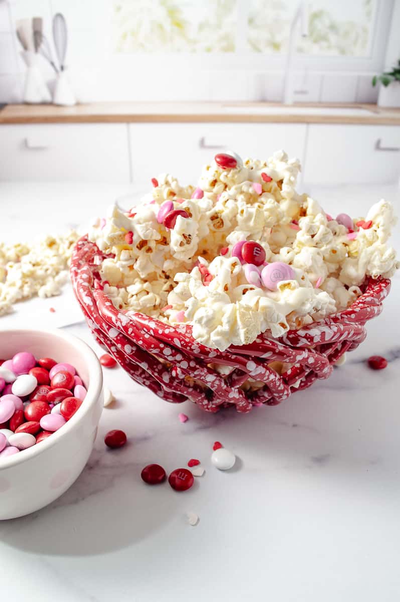 Easy Valentine's White Chocolate M&M Popcorn Treat in a red basket bowl on a white counter in a white kitchen with popcorn on the counter and a small bowl with M and M in it.